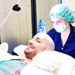 Das Bild zeigt eine Szene in einer modernen medizinischen Klinik. Ein Mann liegt entspannt auf einem Behandlungsstuhl und lächelt zufrieden, während eine Ärztin oder Pflegekraft mit grüner Schutzhaube und Maske eine Haartransplantation durchführt. Der Mann hebt den Daumen hoch, was darauf hinweist, dass er mit dem Verlauf der Behandlung zufrieden ist. Dieses Bild veranschaulicht das Thema Haartransplantation in Wien.