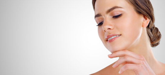 Das Bild zeigt eine junge Frau mit glatter, makelloser Haut und braunen, im Nacken zusammengebundenen Haaren. Sie schaut sanft nach unten und berührt ihr Kinn mit den Fingerspitzen, wodurch der Bereich unter ihrem Kinn – das sogenannte Doppelkinn – betont wird. Die entspannte und gepflegte Ausstrahlung der Frau verleiht dem Bild eine ästhetische und professionelle Note. Diese Darstellung illustriert die Möglichkeit, ein Doppelkinn entfernen mit Spritze zu behandeln. Die Behandlung zur Doppelkinnentfernung mit einer speziellen Injektionsmethode wird bei Dermacare Wien durchgeführt.